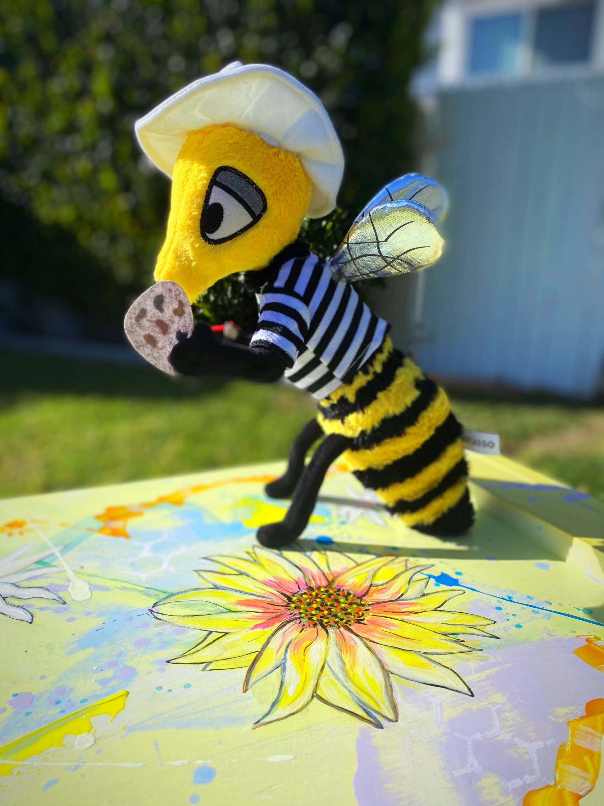 Beecasso plush mascot, a bee in a beret and striped shirt sitting on a painted sunflower